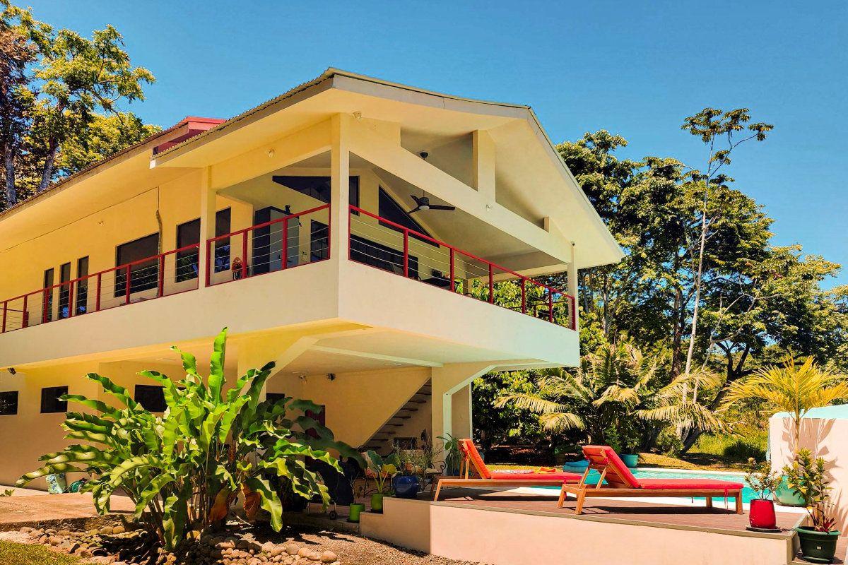 Vista 3/4 de la casa de dos niveles rodeada de árboles y vegetación tropical.